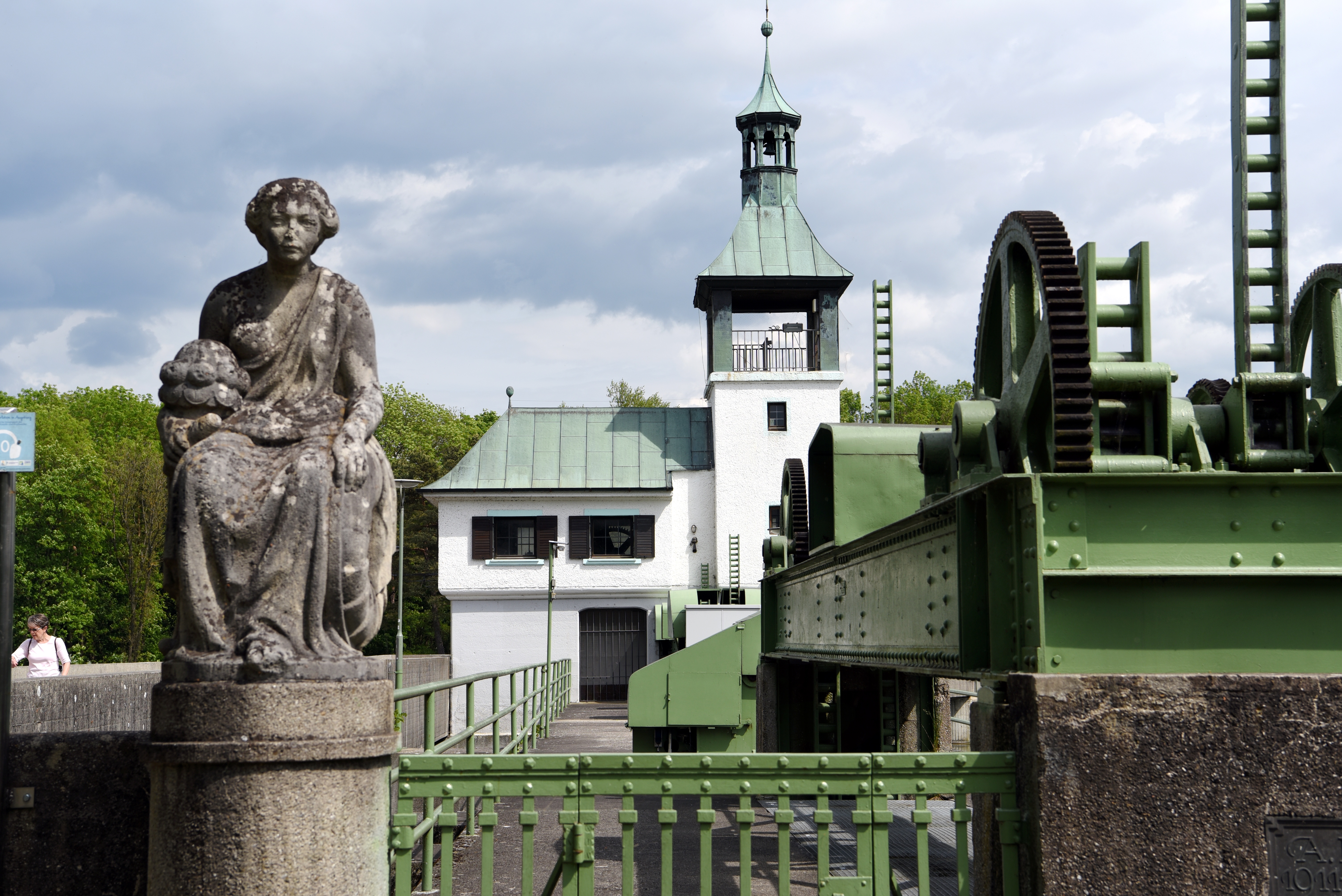 Im Vordergrund eine steinerne Skulptur, daneben eine Schleusen-Anlage. Im Hintergrund ein weißes Haus mit Glockenturm. Alles steht am Rand der Brücke am Hochablass.