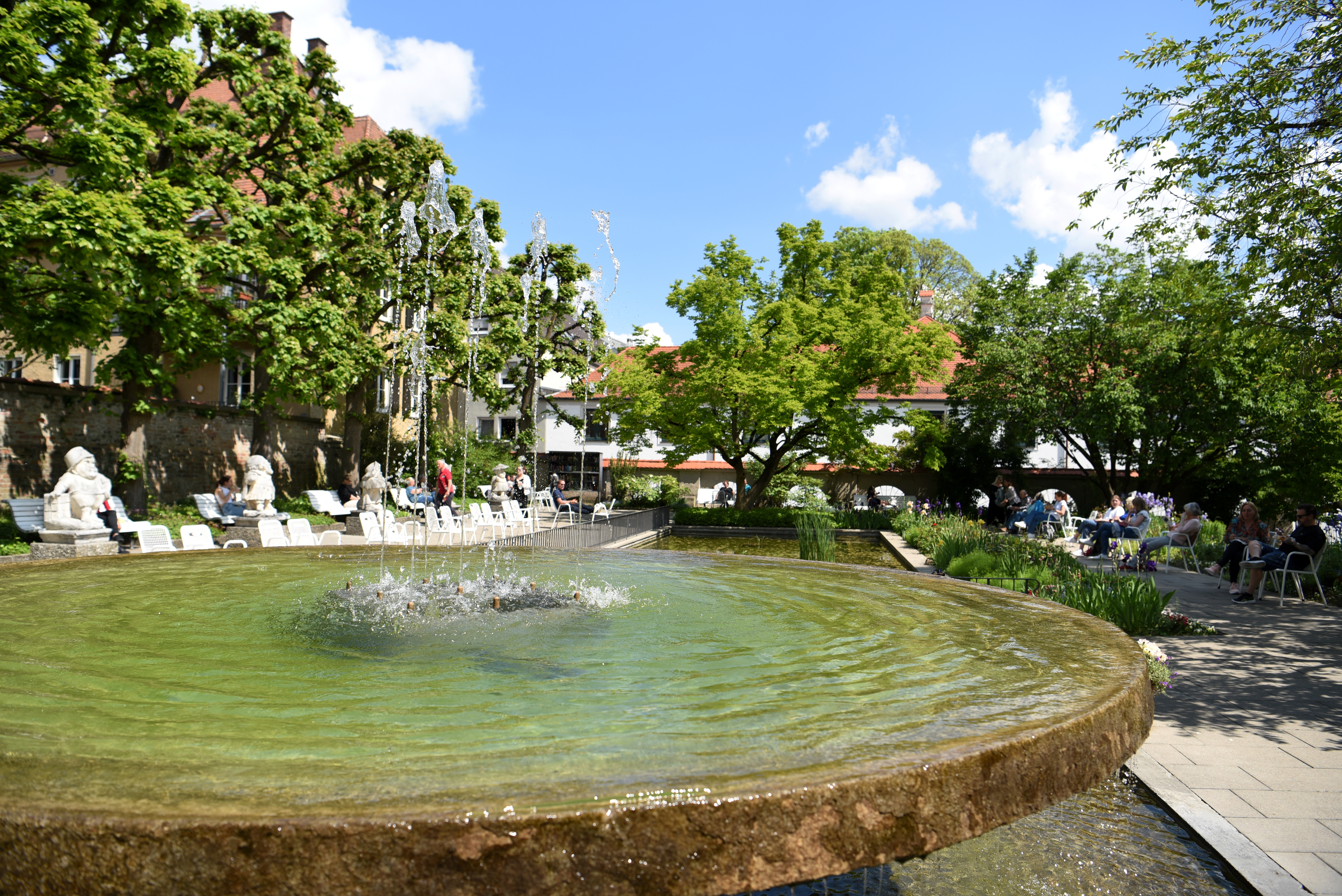 Ein Brunnen in einem großen Garten. Viele Leute liegen auf Liegestühlen