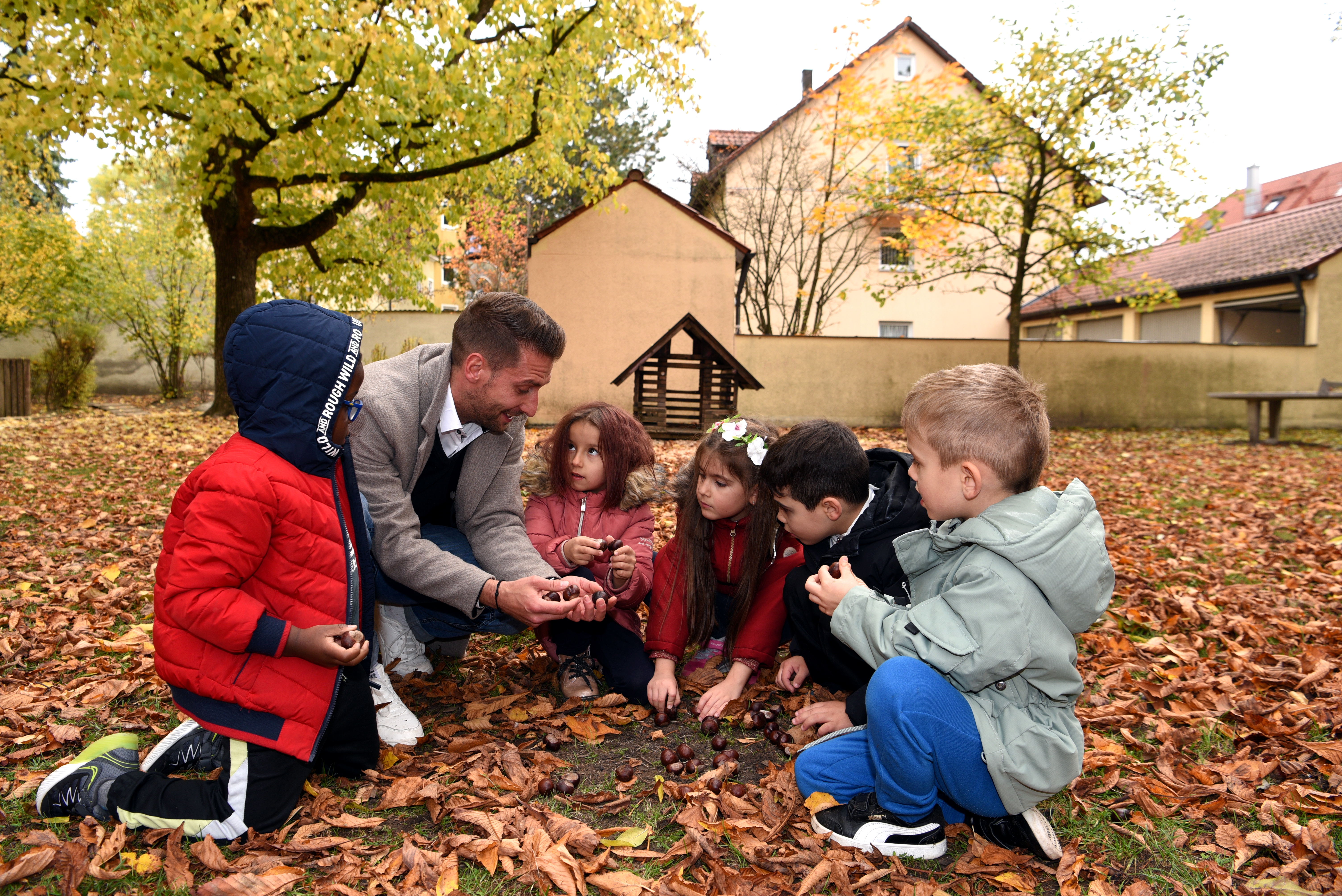 Ein Mann und fünf Kinder knien in einem Garten und spielen mit Herbstlaub und Kastanien.