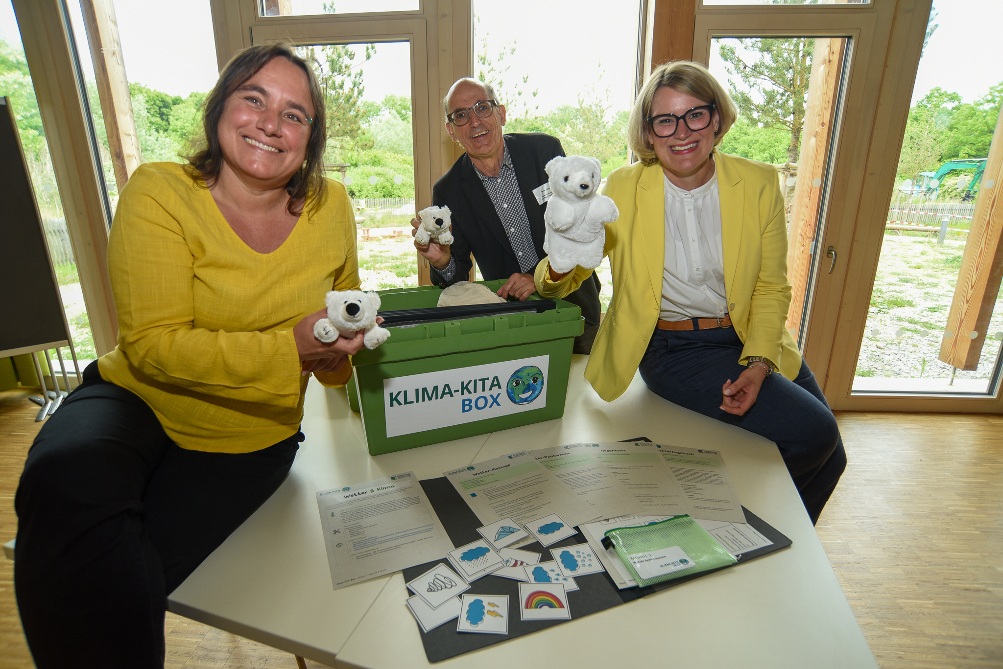 zwei Frauen und ein Mann lächeln in die Kamera, zwischen ihnen eine Plastikbox mit der Aufschrift Klima-Kita-Box