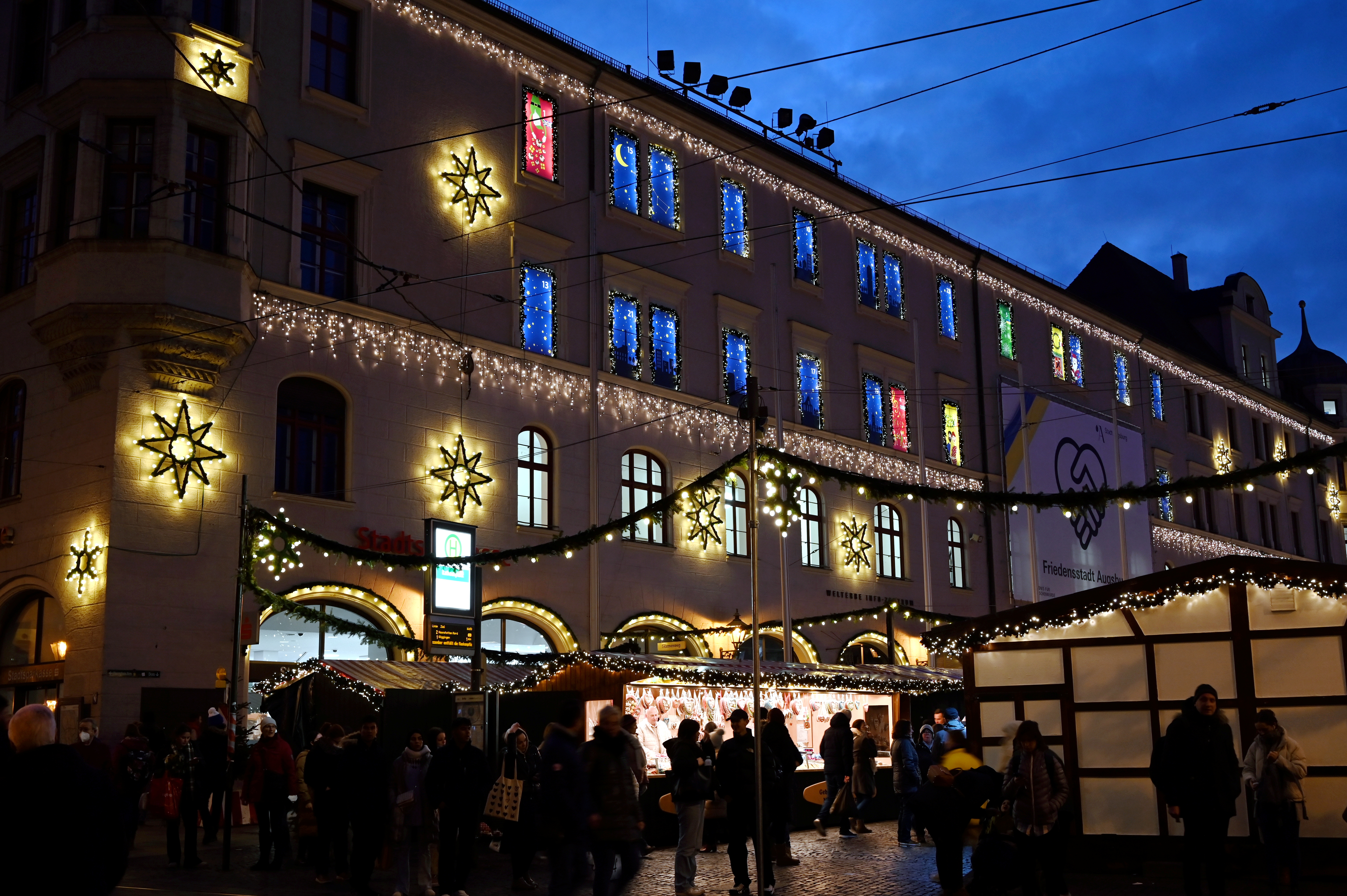 Das Verwaltungsgebäude am Rathausplatz, festlich beleuchtet mit Weihnachtssternen. Im Vordergrund ist der Christkindlesmarkt zu sehen.