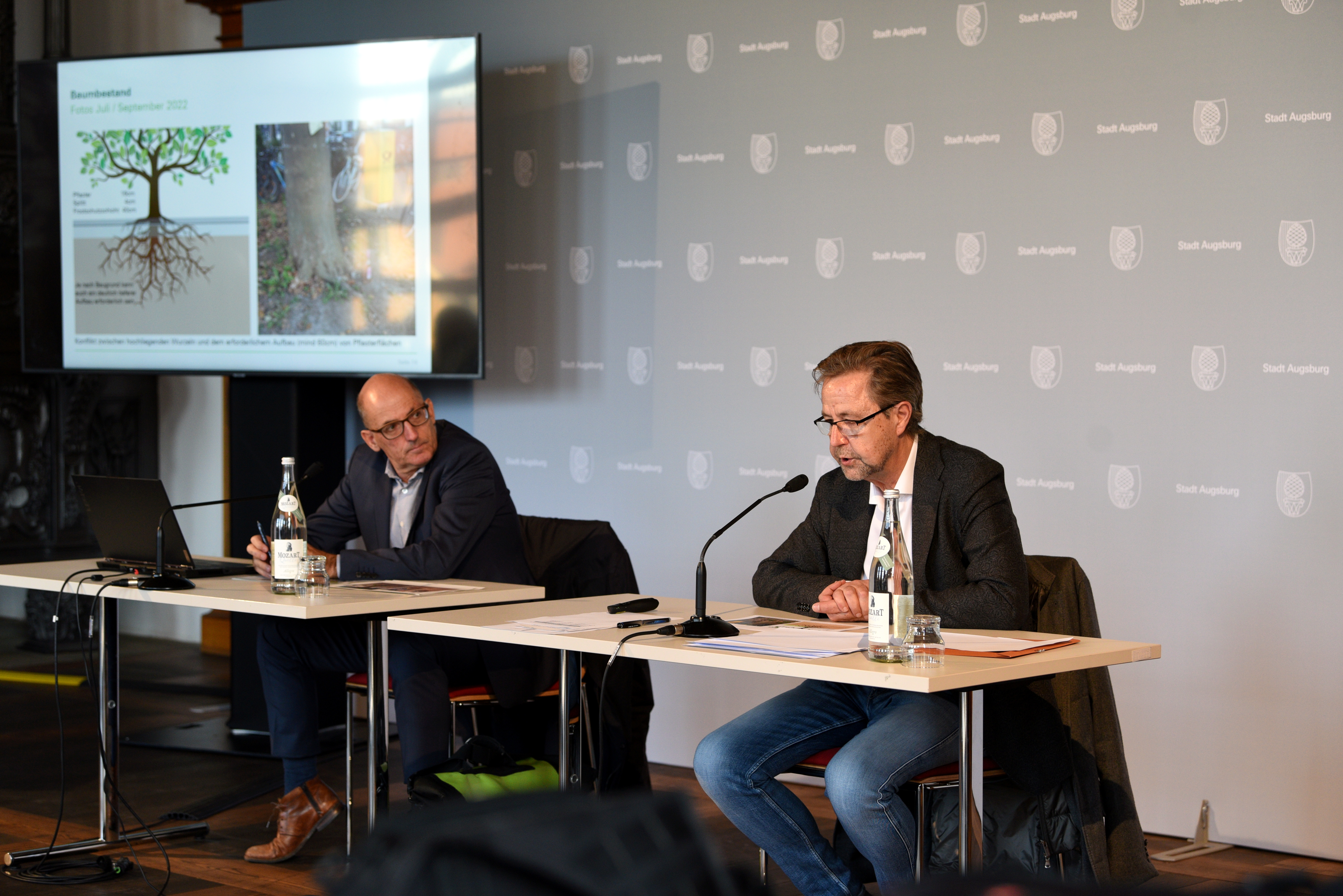 Umweltreferent Reiner Erben und Baureferent Gerd Merkle sitzen an einem Tisch und sprechen zu Journalisten. Im Hintergrund ist ein Bildschirm zu sehen. Auf diesem wird erklärt, wie viel Platz ein Baum zum Wurzeln braucht. 