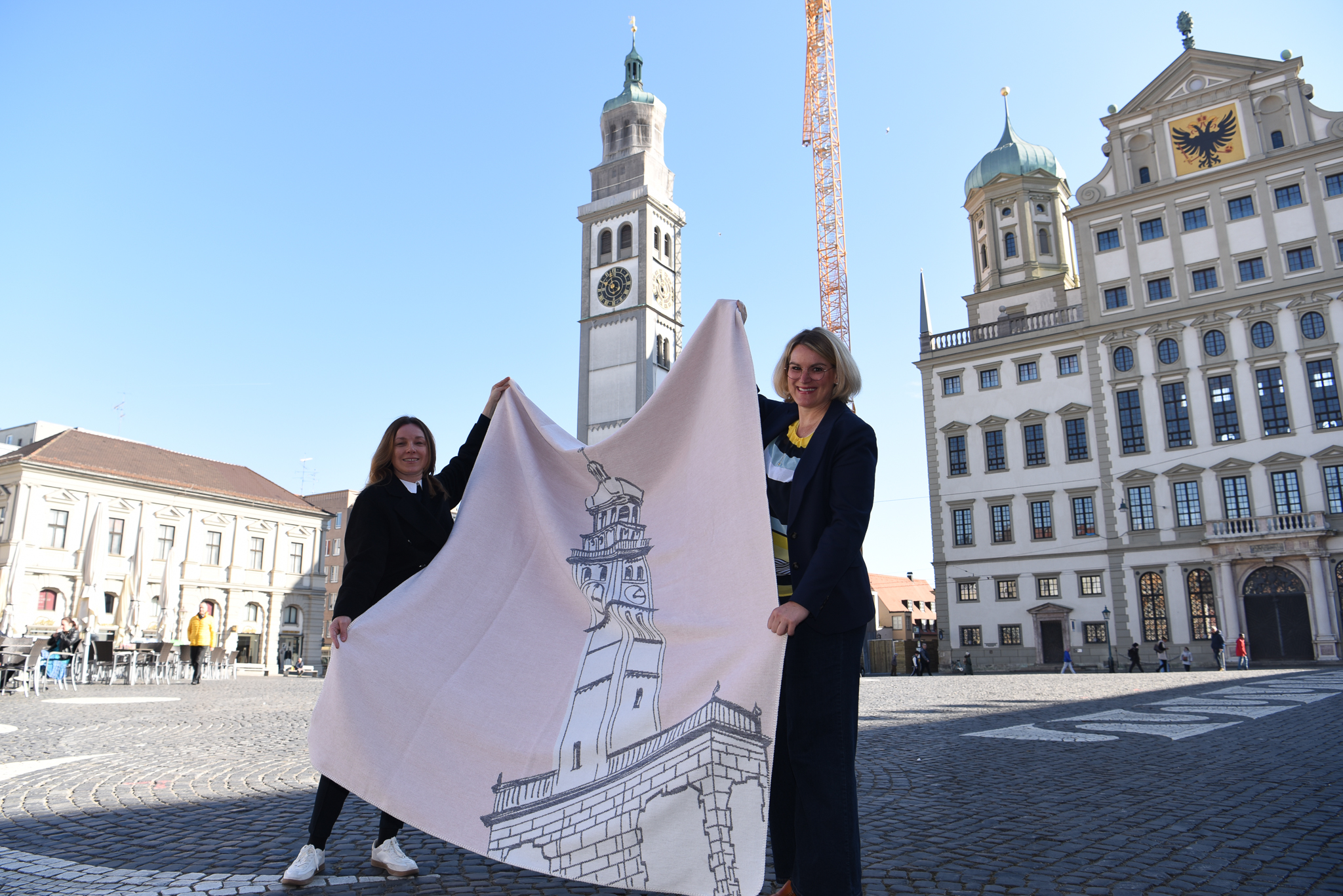 zwei Frauen halten vor dem Perlachturm eine Decke in die Höhe, auf der der Perlachturm abgebildet ist