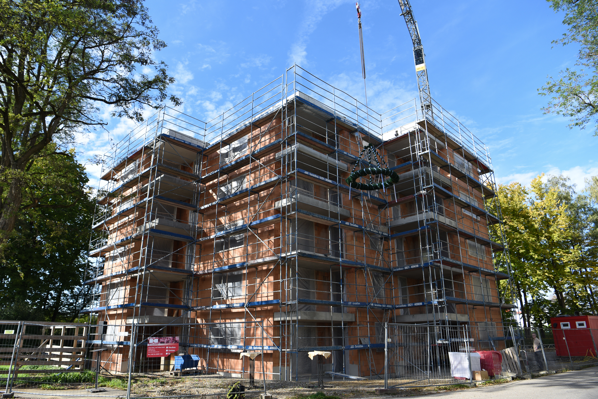 mehrstöckiger Rohbau mit Gerüst, rechts daneben schwebt die Richtkrone an einem Kran