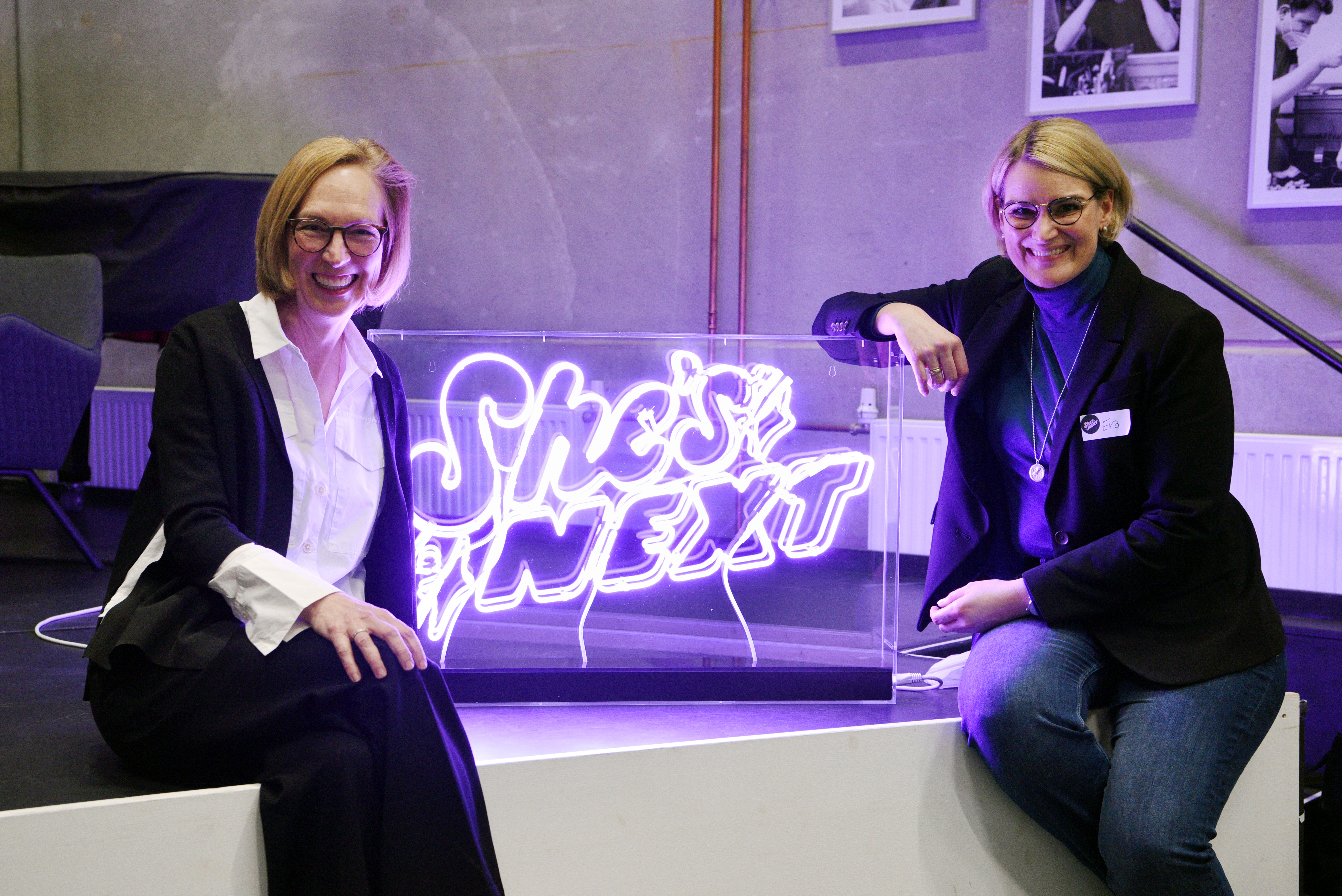Zwei Frauen sitzen neben einer Leuchtschrift mit dem Text "She's next". Rechts im Bild ist Oberbürgermeisterin Eva Weber