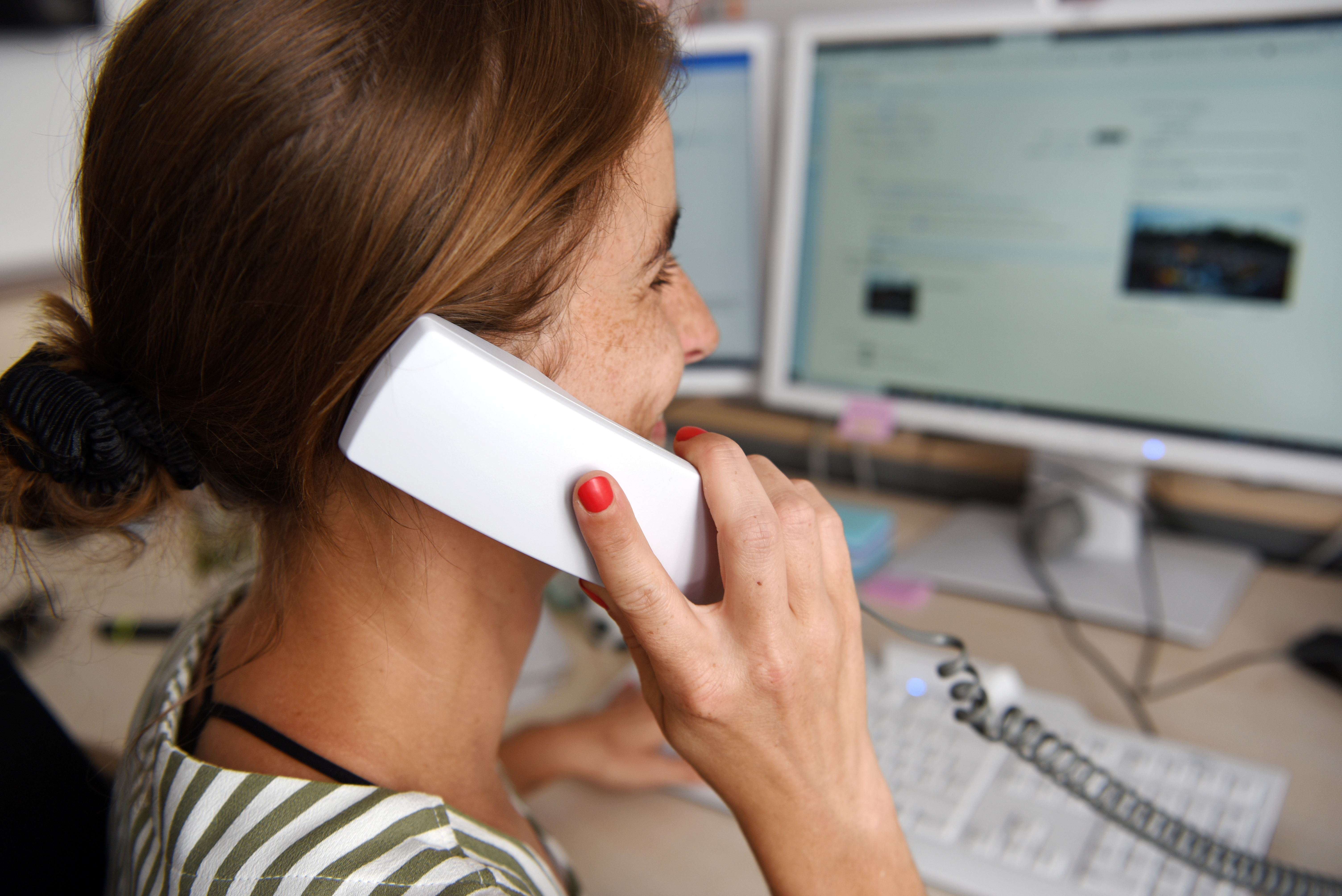 Eine Frau sitzt an einem Computer. Sie hat einen Telefonhörer in der Hand und hält ihn an ihr rechtes Ohr.