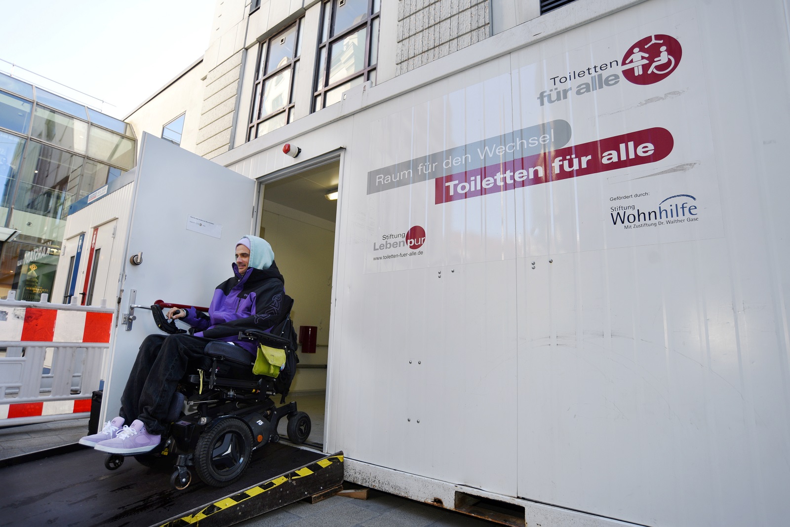 Ein Mann im Rollstuhl fährt über eine Rampe aus einem Toiletten-Container