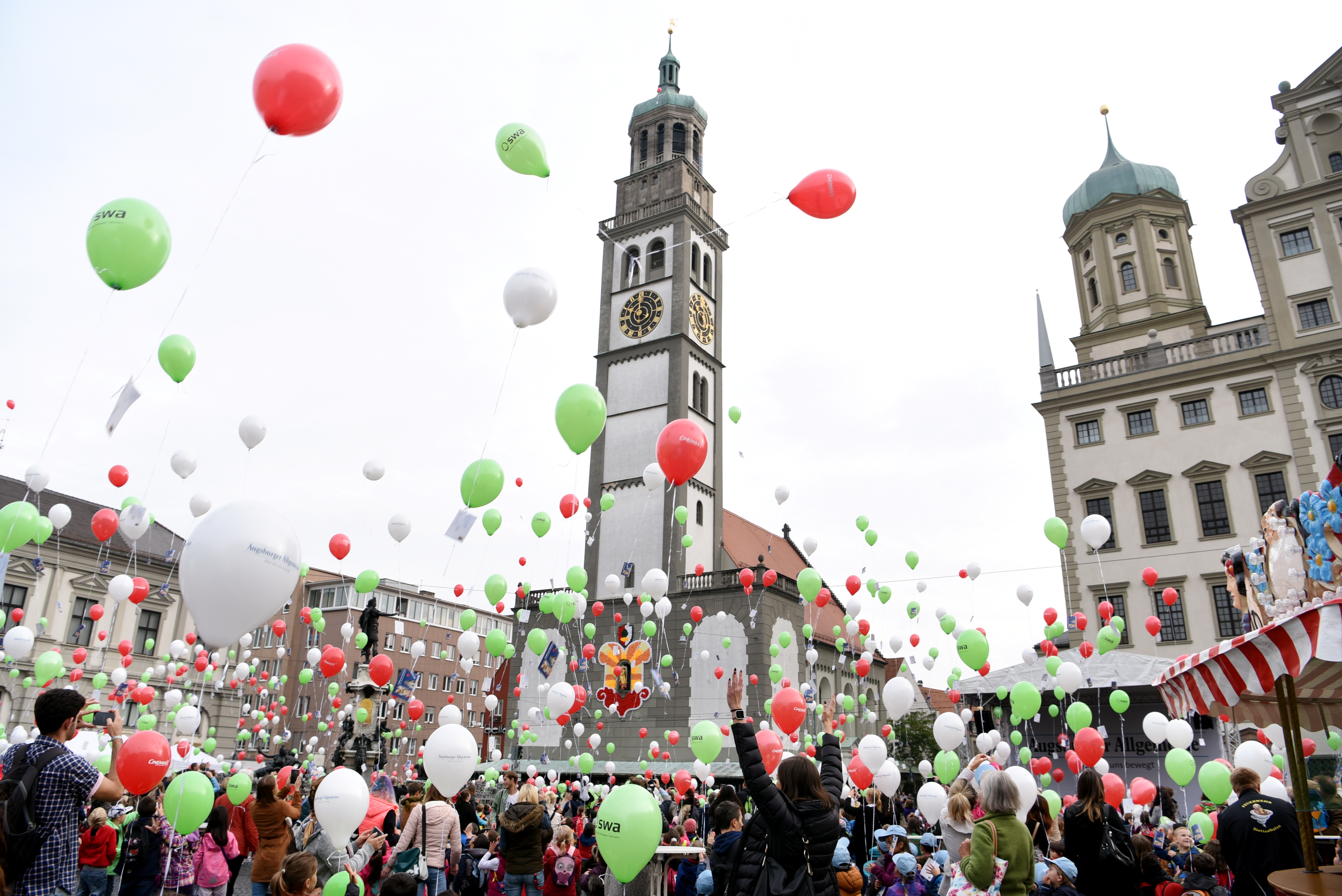 Hunderte Kinder stehen auf dem Rathausplatz, während das Turamichele in einem Fenster des Rathauses zu sehen ist. Die Kinder lassen Luftballons in den Farben Rot, Grün und Weiß steigen.
