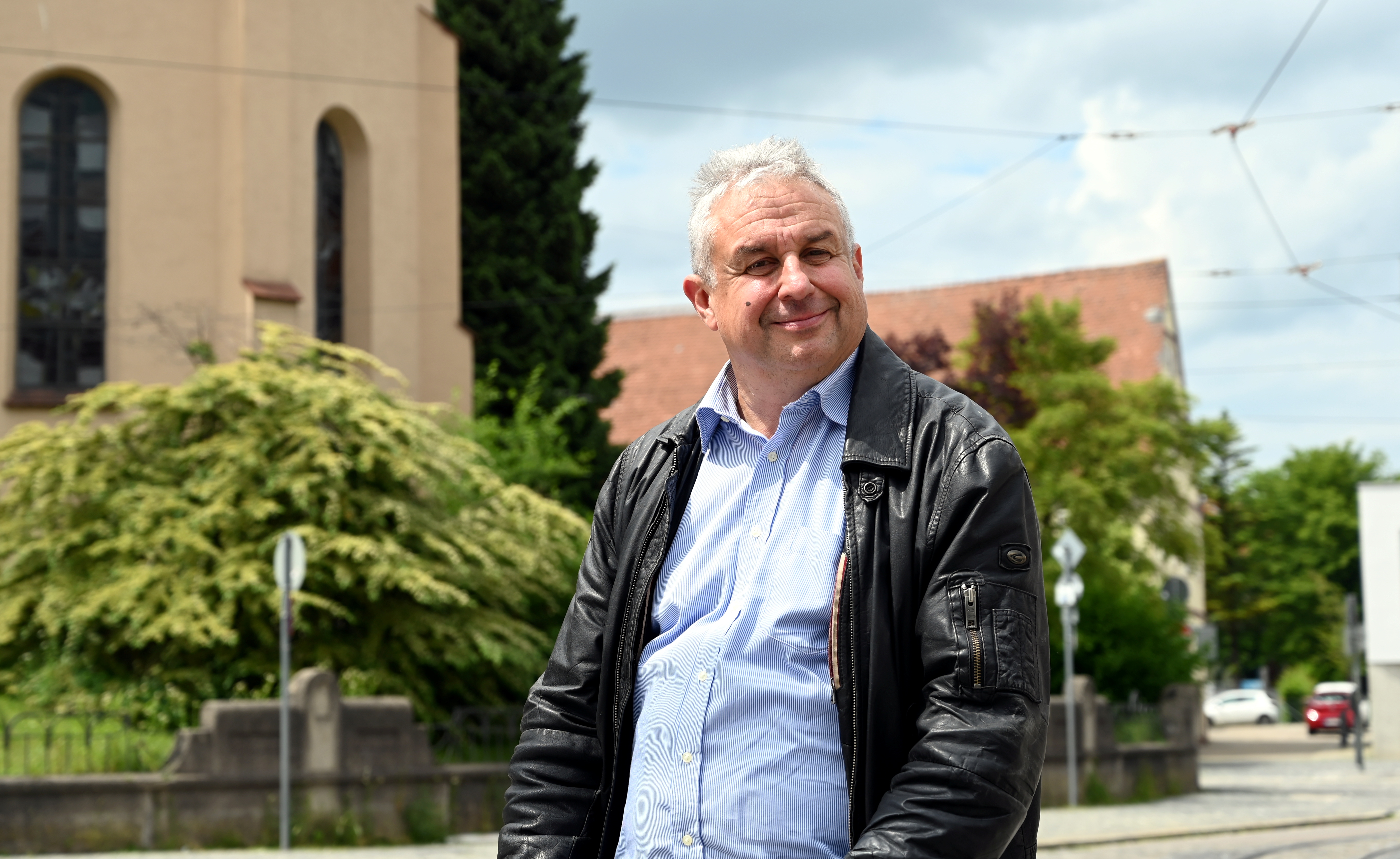 Ein Mann in Hemd und Lederjacke steht vor einer Kirche.