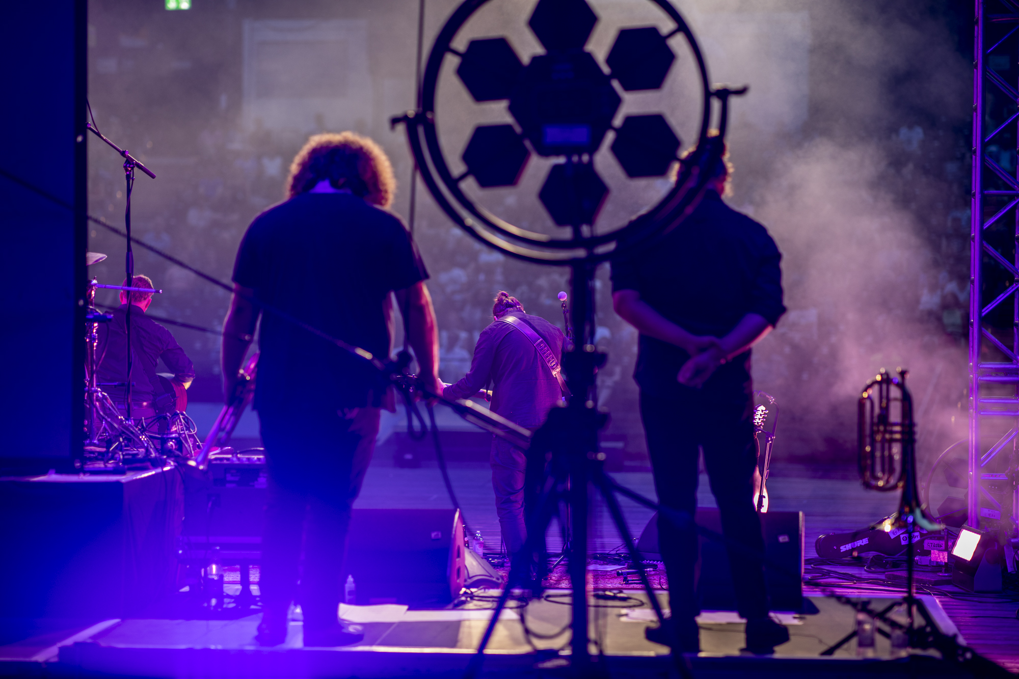 Es ist eine Band auf der Bühne von hinten zu sehen. Das Licht ist in violetten Tönen gehalten, man sieht Nebel von einer Nebelmaschine.
