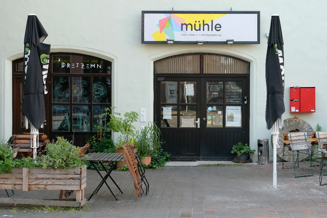 Frontalfoto des Eingangs der Kresslesmühle. Über der großen hölzernen Eingangstüre hängt ein Schild mit dem Namen. Links und rechts sieht man Tische, Stühle und Schirme des Restaurants dreizehn. 