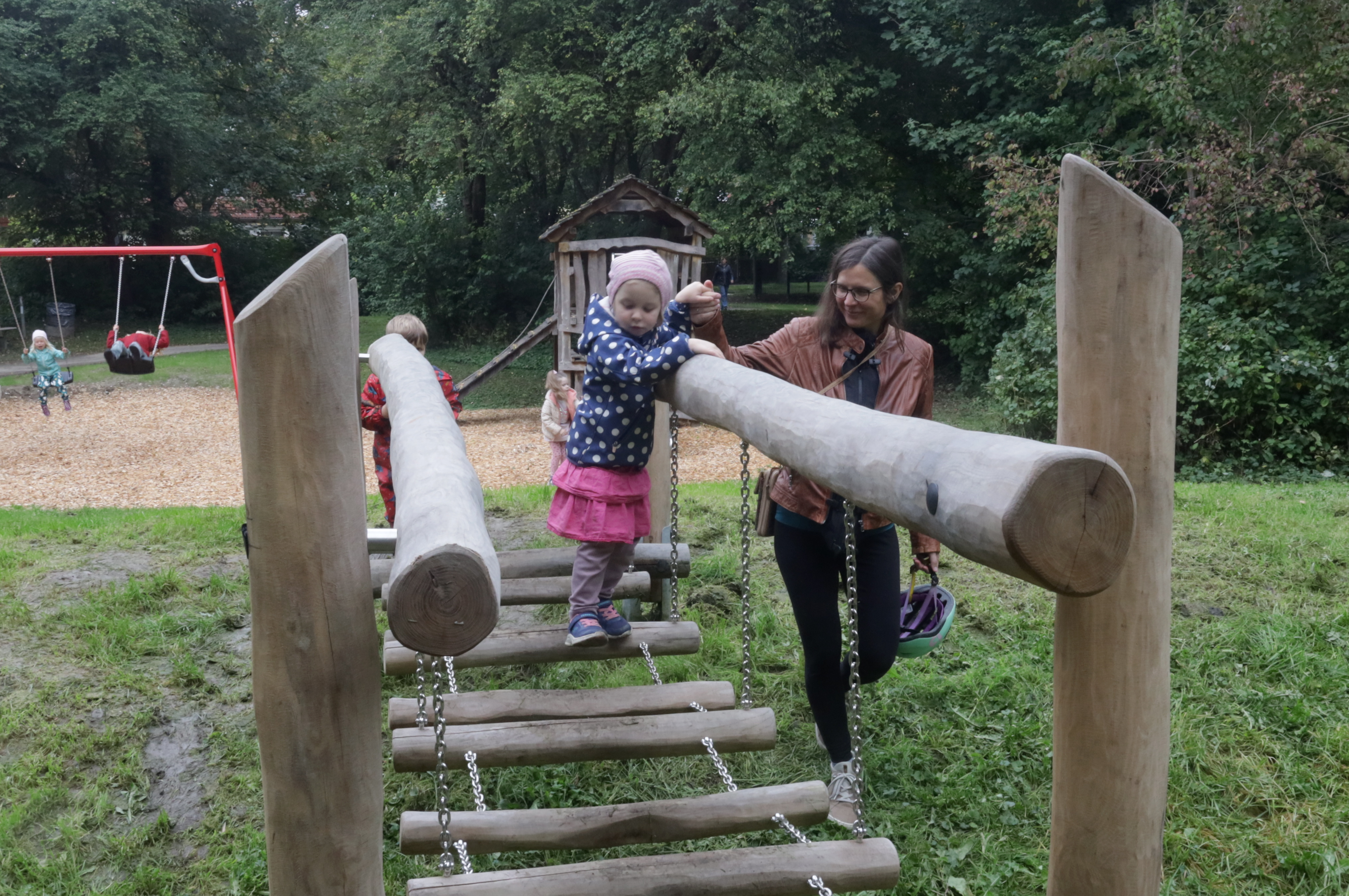 Ein kleines Mädchen balanciert über eine Wackelbrücke. Ihre Mutter geht nebenher und hält ihr eine Hand