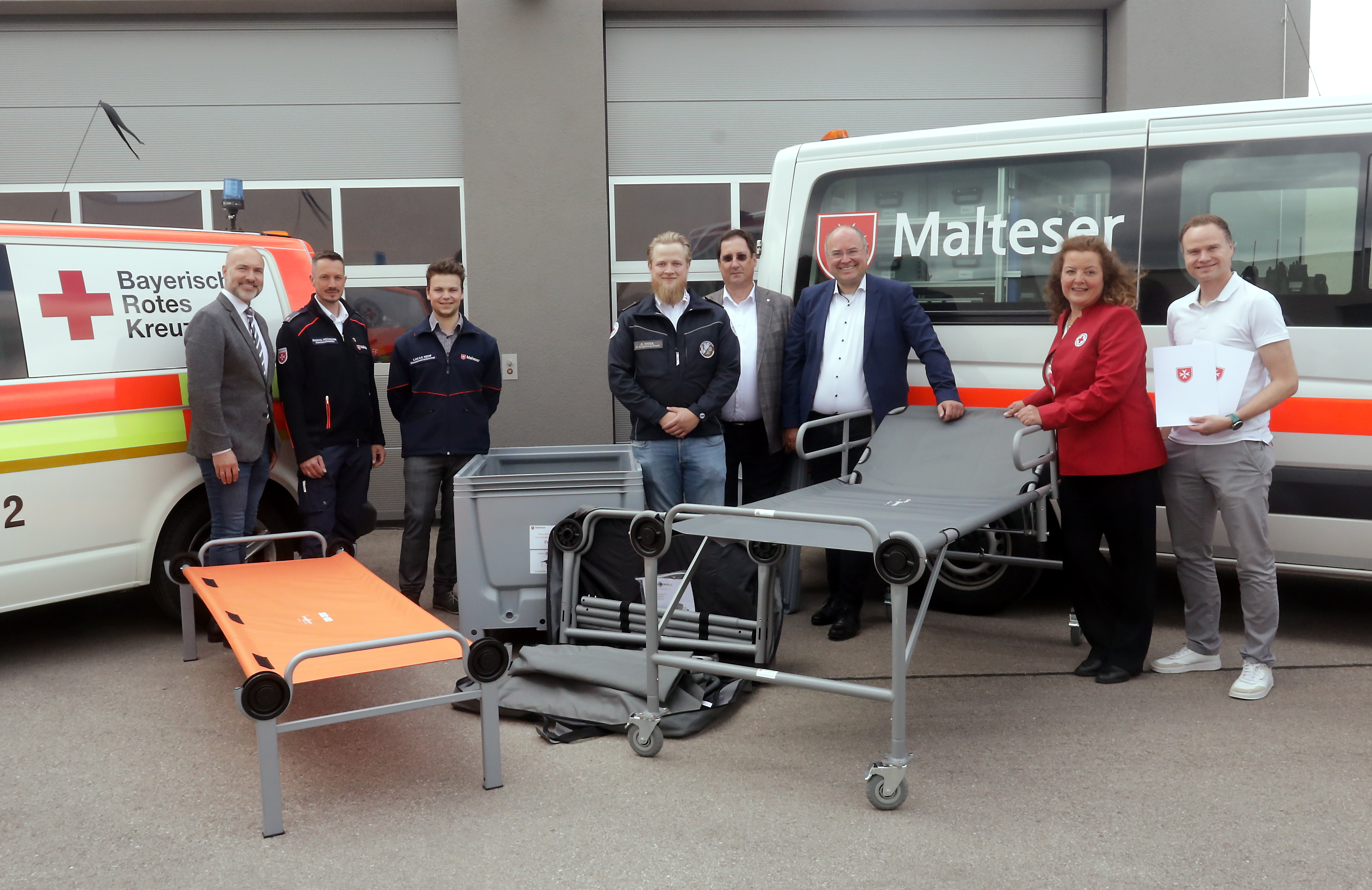 Acht Menschen stehen neben zwei Pflegebetten. Im Hintergrund Rettungsfahrzeuge vom Roten Kreuz und vom Maltester Hilfsdienst