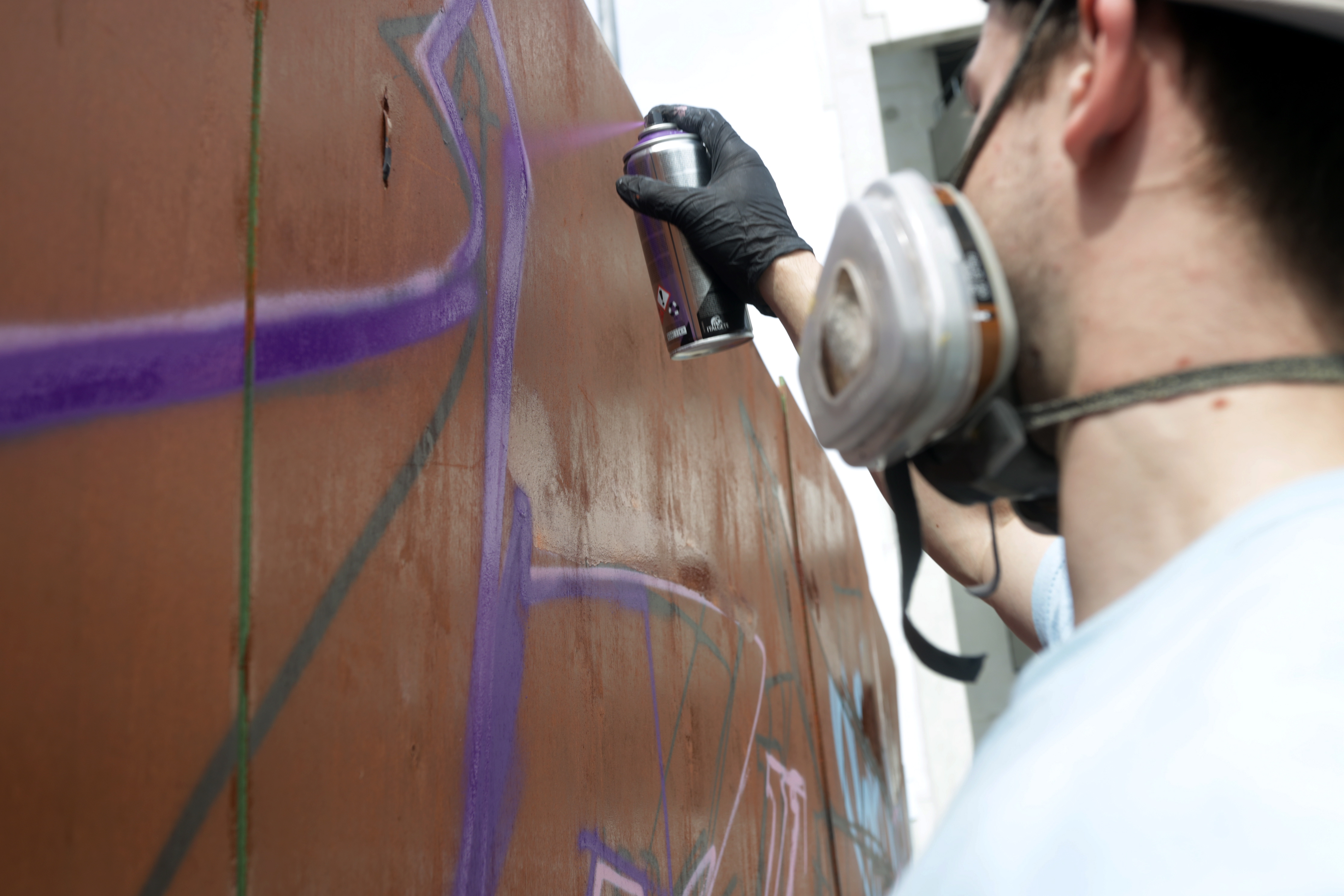 Ein Graffiti-Künstler mit Atemmaske sprayt auf eine braune Wand.