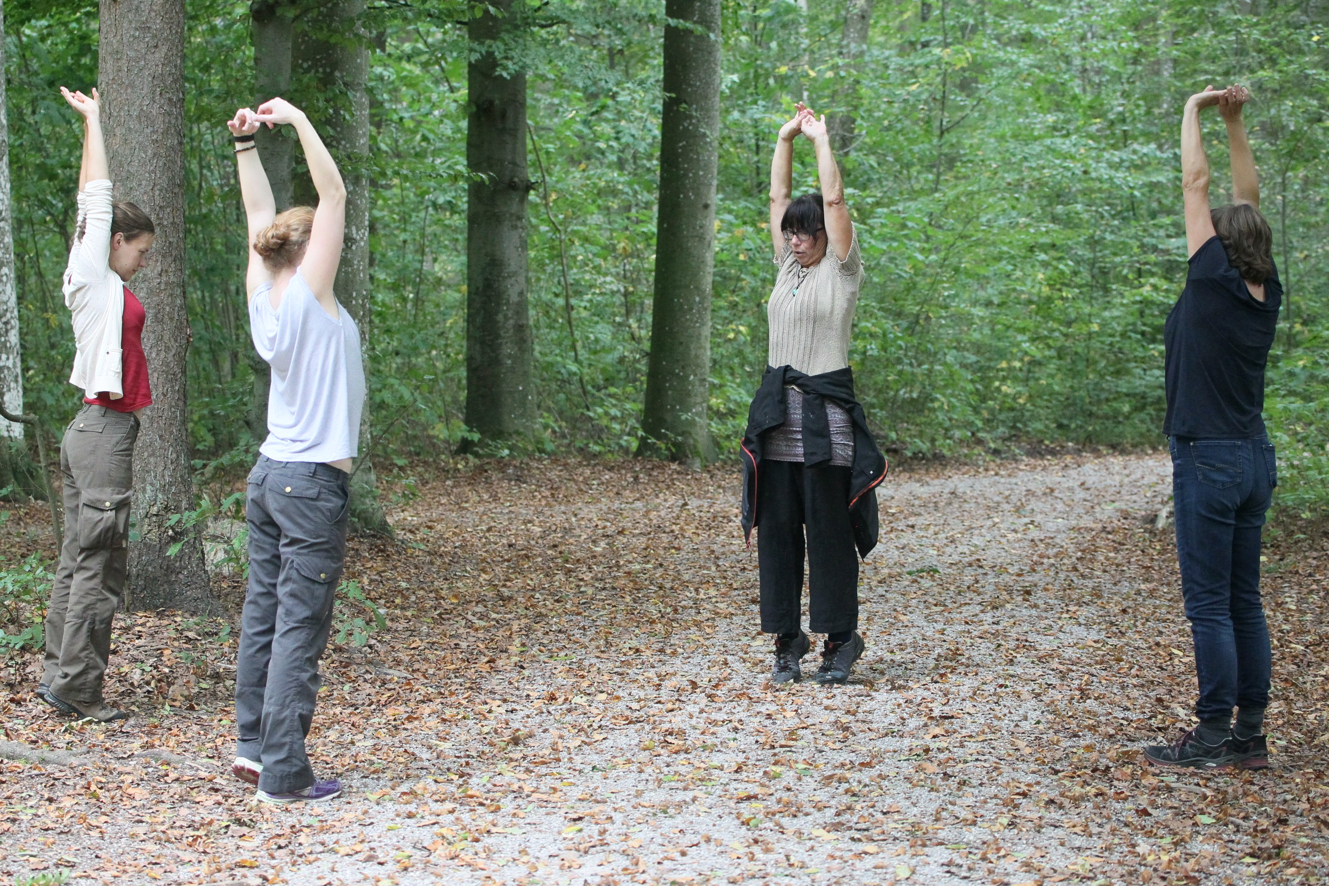 Vier Frauen stehen auf einem Weg im Wald. Sie strecken ihre Arme nach oben.