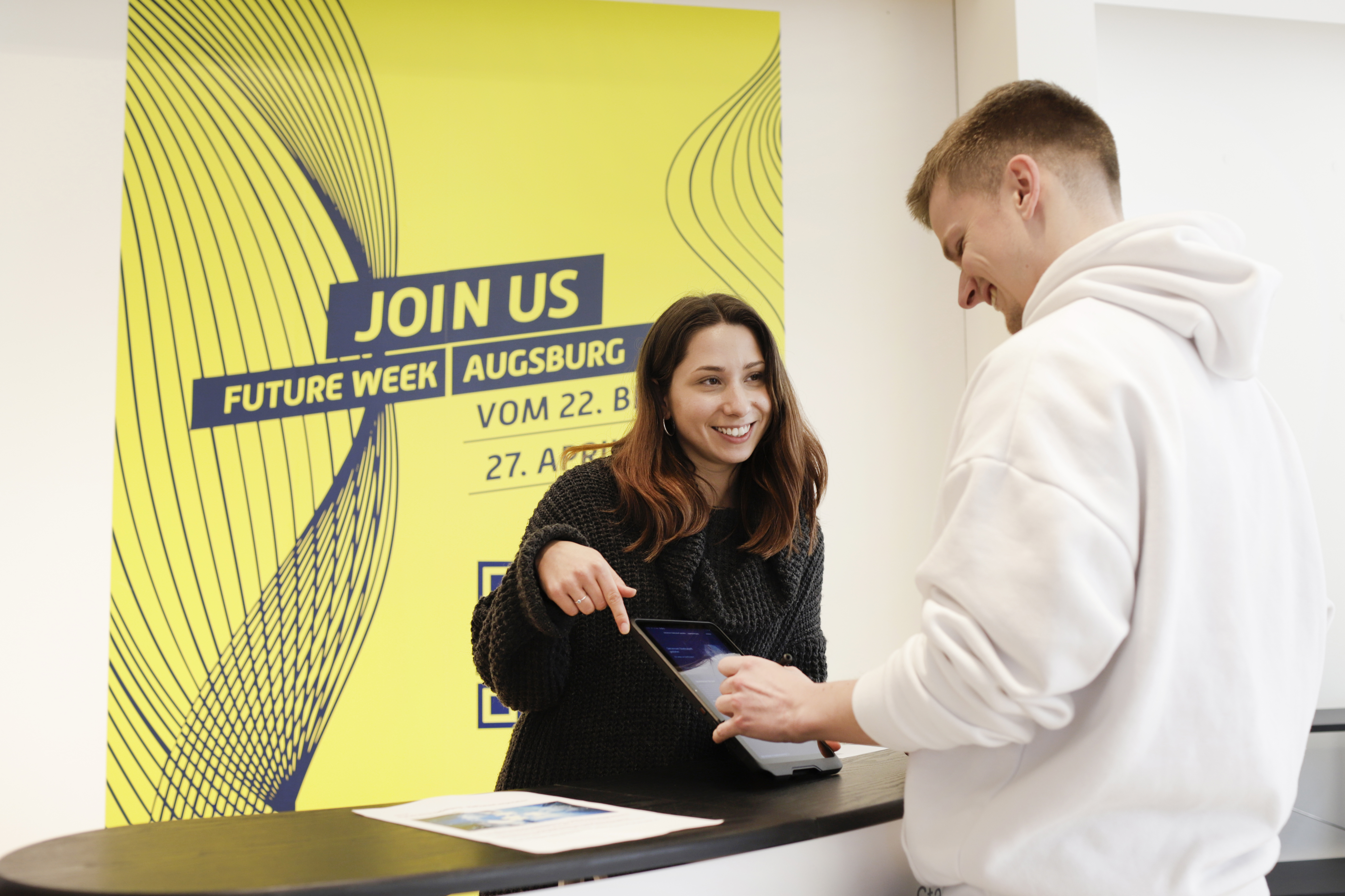 Eine Frau berät einen Mann. Im Hintergrund hängt ein Plakat zur "Future Week Augsburg"
